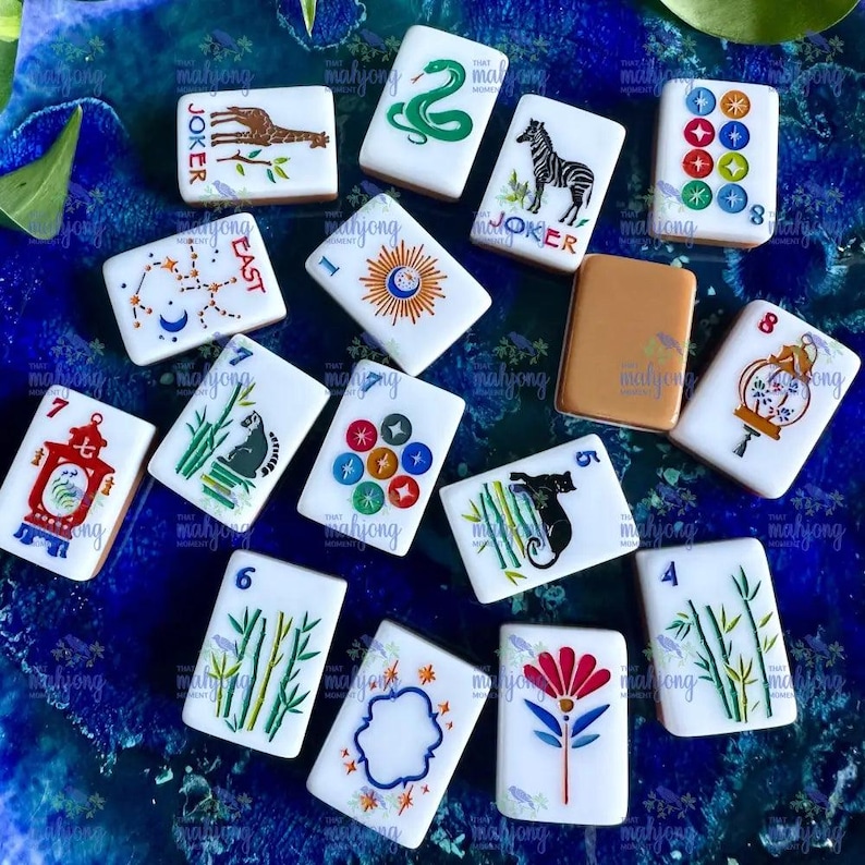 May include: A collection of white Mahjong tiles with various colorful illustrations, including a zebra, a snake, and a giraffe. Some tiles feature numbers and symbols. The tiles are arranged on a blue surface.