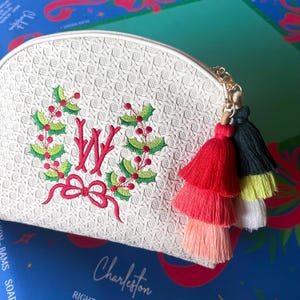 K&ouml;nnte beinhalten: Wei&szlig;es, strukturiertes Kosmetikt&auml;schchen mit goldenem Rei&szlig;verschluss und farbenfrohem Quastenanh&auml;nger. Die Tasche hat einen gestickten Kranz mit roter Schleife und dem Buchstaben "W". Die Quasten sind rot, rosa, schwarz, gelb und wei&szlig;.
