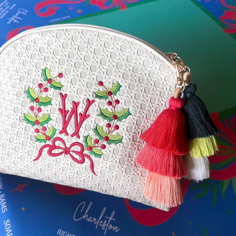 K&ouml;nnte beinhalten: Wei&szlig;es, strukturiertes Kosmetikt&auml;schchen mit goldenem Rei&szlig;verschluss und farbenfrohem Quastenanh&auml;nger. Die Tasche hat einen gestickten Kranz mit roter Schleife und dem Buchstaben "W". Die Quasten sind rot, rosa, schwarz, gelb und wei&szlig;.