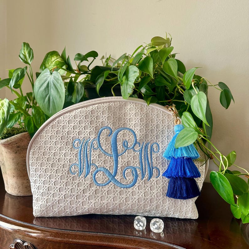K&ouml;nnte beinhalten: Eine beige Kosmetiktasche mit Webmuster und blauem gesticktem Monogramm. Die Tasche hat einen goldenen Rei&szlig;verschluss mit einem blauen Quastenanh&auml;nger. Die Tasche steht auf einer dunklen Holzoberfl&auml;che mit Pflanzen im Hintergrund.