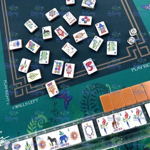 May include: A Mahjong game set with white tiles featuring various symbols and characters. The game is set on a green mat with floral patterns and text that reads "PLAY RIGHT" and "WALLS LEFT". Includes dice and a rack of tiles.