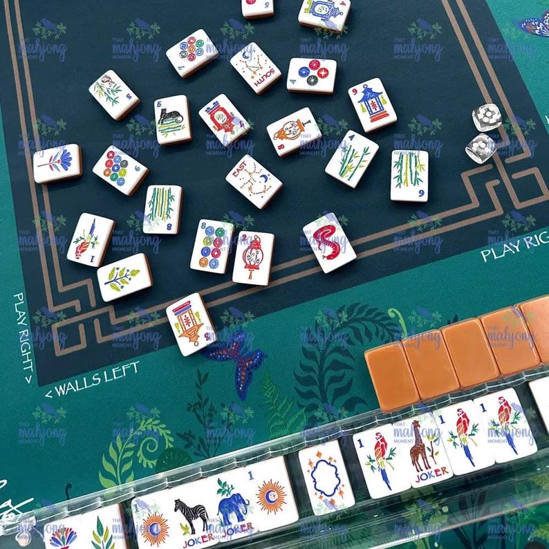May include: A Mahjong game set with white tiles featuring various symbols and characters. The game is set on a green mat with floral patterns and text that reads "PLAY RIGHT" and "WALLS LEFT". Includes dice and a rack of tiles.