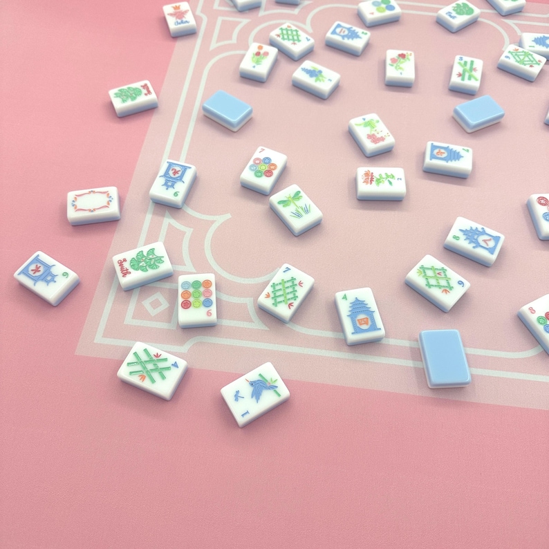 May include: A collection of small, rectangular Mahjong tiles scattered on a pink surface. The tiles are white with various colorful symbols and designs, including flowers, numbers, and geometric patterns. Some tiles are solid light blue.