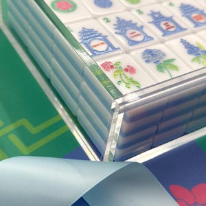 May include: A close-up of a Mahjong set with white tiles featuring colorful illustrations and numbers. The tiles are in a clear acrylic case. The background includes a blue ribbon and a patterned surface.