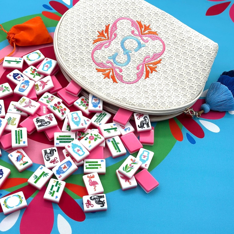 K&ouml;nnte beinhalten: Ein wei&szlig;es Mahjong-Set im Rattan-Stil mit einer passenden Rei&szlig;verschlusstasche mit einem gestickten Blumenmuster und dem Buchstaben "S". Das Set enth&auml;lt rosa Spielsteine und einen orangefarbenen Kordelzugbeutel, alles auf einer farbenfrohen, floral gemusterten Oberfl&auml;che.