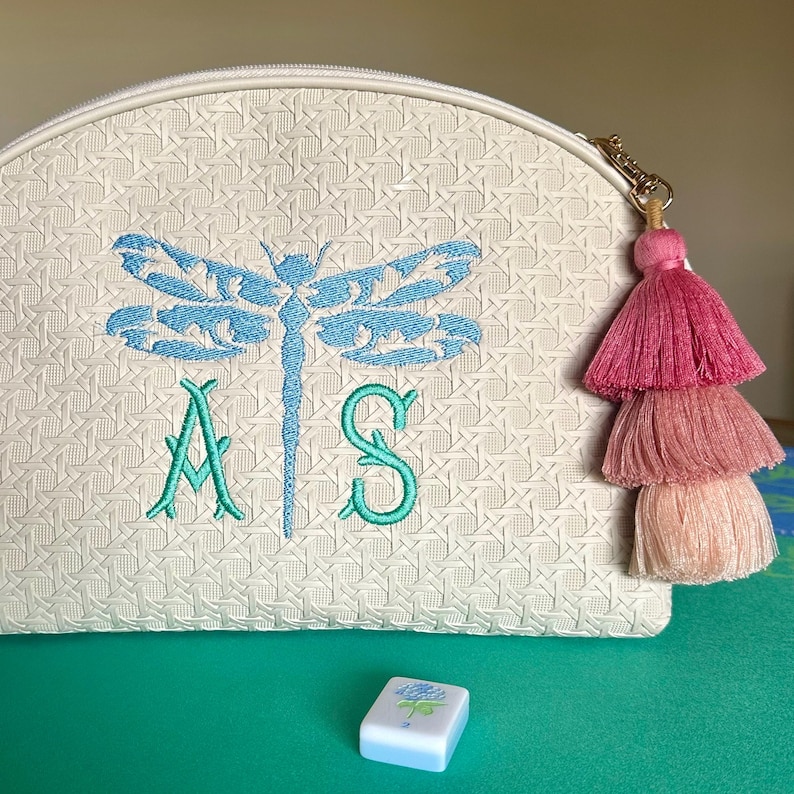 K&ouml;nnte beinhalten: Cremefarbene Kosmetiktasche mit einem Libellen- und Initialen-Stickmuster in T&uuml;rkis und Gr&uuml;n. Die Tasche hat einen goldenen Rei&szlig;verschluss mit einem rosa-, pfirsich- und cremefarbenen Quastenanh&auml;nger. Eine kleine Kachel mit floralem Design ist im Vordergrund.