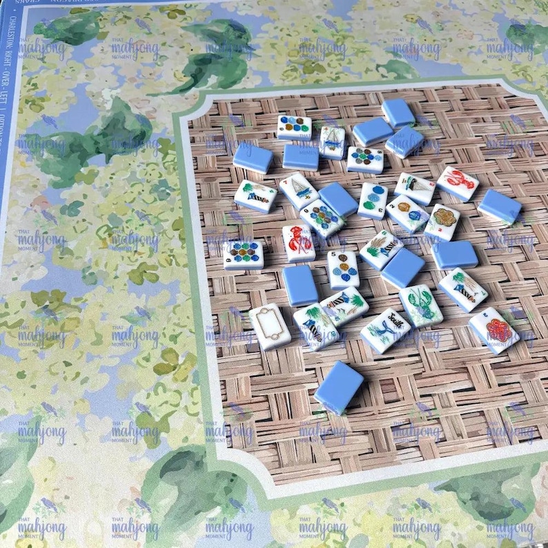 May include: A mahjong game set with tiles featuring various illustrations, including a lobster, sailboat, and flowers. The tiles are white with blue backs, scattered on a woven bamboo-style game board. The board has a floral design.