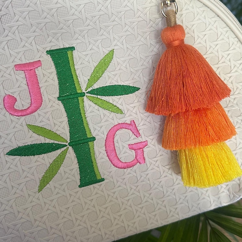 K&ouml;nnte beinhalten: Wei&szlig;e Tasche mit Rei&szlig;verschluss und Bambus- und Monogramm-Design. Die Tasche hat die gestickten Buchstaben "J" und "G" in Rosa und einen gr&uuml;nen Bambusstamm mit gr&uuml;nen Bl&auml;ttern. Ein dreifarbiger Quastenanh&auml;nger in Orange, Gelb und Orange h&auml;ngt am Rei&szlig;verschluss.