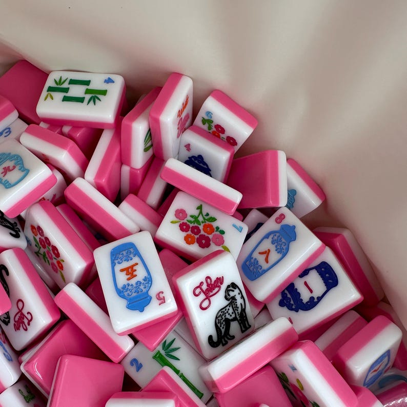 May include: A collection of pink and white Mahjong tiles. The tiles feature various symbols, numbers, and illustrations, including flowers, bamboo, and a leopard. The tiles are stacked together, creating a colorful and engaging display.
