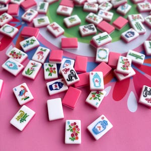 May include: A collection of pink and white Mahjong tiles with various colorful illustrations and characters. The tiles are scattered on a pink surface with a green and white patterned background. Some tiles have floral designs, while others feature birds and other symbols.