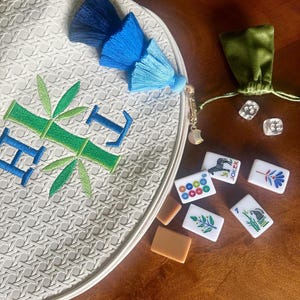 K&ouml;nnte beinhalten: Eine wei&szlig;e, runde, gewebte Tasche mit blau und gr&uuml;n gesticktem Bambus und den Buchstaben "HI". Die Tasche hat eine blaue Quaste und einen goldenen Anh&auml;nger. Mahjong-Steine und ein gr&uuml;nes Samtbeutel liegen neben der Tasche.