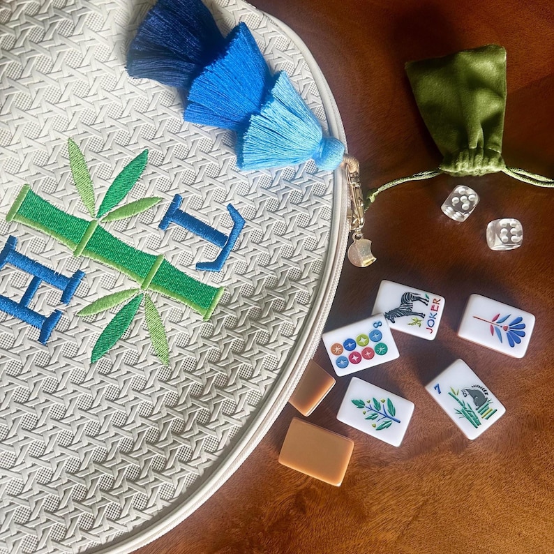 K&ouml;nnte beinhalten: Eine wei&szlig;e, runde, gewebte Tasche mit blau und gr&uuml;n gesticktem Bambus und den Buchstaben "HI". Die Tasche hat eine blaue Quaste und einen goldenen Anh&auml;nger. Mahjong-Steine und ein gr&uuml;nes Samtbeutel liegen neben der Tasche.