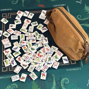 May include: A collection of white Mahjong tiles with colorful illustrations of butterflies, plants, and compasses scattered on a green surface. A tan canvas bag with a zipper and leather accents sits nearby. The game board has text that reads "PLAY RIGHT" and "WALLS LEFT".