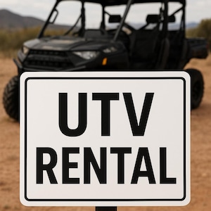 May include: A black UTV (Utility Task Vehicle) is in the background, with a sign in the foreground that reads "UTV RENTAL" in bold, black capital letters. The sign is white with a black border, and the background is a desert landscape.
