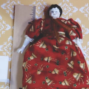 May include: A vintage porcelain doll with a painted face, black hair, and a red dress adorned with gold bells and green stars. The doll's dress has white lace trim. The doll's legs are white porcelain with black boots. The doll is approximately 6 inches tall.