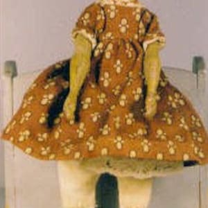 May include: A vintage wooden doll with painted features, wearing a brown and white floral dress and white bloomers. The doll is sitting on a small wooden chair.