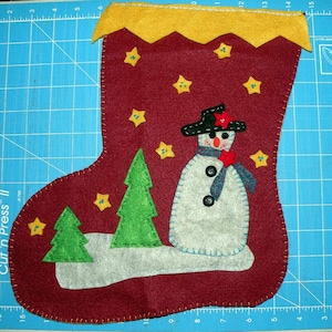 Puede incluir: Una media de fieltro roja con un puño de fieltro amarillo y un muñeco de nieve de fieltro blanco con un sombrero negro y una bufanda roja. La media está decorada con estrellas de fieltro amarillo y un árbol de Navidad de fieltro verde.