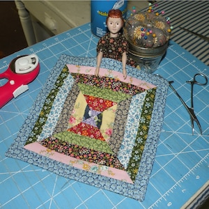 May include: A colorful patchwork quilt square with a blue, pink, and white border. The square is made of various fabrics with floral patterns and is displayed on a blue cutting mat.