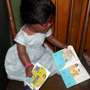 May include: A young person wearing a light blue dress and glasses is sitting on a wooden chair. They are holding a book with the title "Three Little Piglets" and a paper doll set. The paper doll set includes a doll with a pink dress and a doll with a yellow dress.