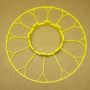 May include: A yellow plastic bird feeder with a circular base and a center ring. The feeder has a decorative design with multiple spokes radiating from the center.