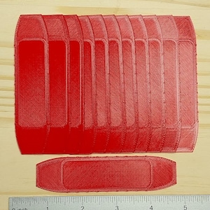 May include: Two red 3D-printed objects are displayed on a wooden surface. The larger object is rectangular with a series of curved vertical sections. The smaller object is a matching red, rectangular shape. A ruler is placed below the objects, showing measurements in inches.
