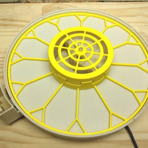 May include: A yellow and off-white plastic incubator tray with a circular design. The tray features a central yellow component with a spiral pattern and a surrounding grid-like structure. A small, off-white component is attached to the side.