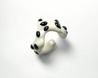 May include: A white ceramic ring with black polka dots. The ring has a curved, open design and is set against a white background. The black dots are evenly spaced across the surface of the ring.