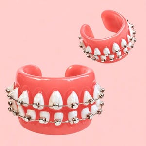 May include: Two cuff bracelets shaped like pink gums with white teeth and silver-colored metal orthodontic braces. The design features a row of teeth with metallic brackets and wires running horizontally across the front of each piece.