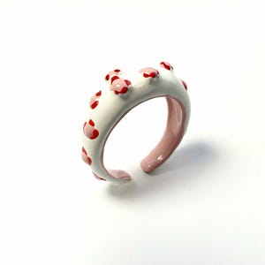 May include: A white ceramic ring with red and pink floral accents. The ring has a smooth, glossy finish and a unique open design. The interior of the ring is a soft pink color.
