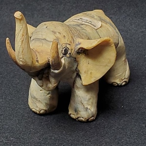 May include: A small, hand-painted ceramic elephant figurine with a textured surface and a light brown and gray color scheme. The elephant has a raised trunk and a tag on its back that reads "Hand Painted Philippines".