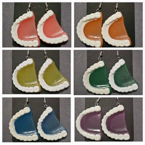 May include: Six pairs of earrings shaped like dentures in various colours, including pink, orange, yellow, green, blue, and purple. Each earring features a white plastic design resembling teeth.