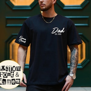 May include: Black t-shirt with the word "Dad" and the year "2025" printed in white. The sleeve has the names "Emma Jacob" printed in white. A circular graphic at the bottom says "CUSTOM Font & Names".