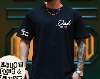 May include: Black t-shirt with the word "Dad" and the year "2025" printed in white. The sleeve has the names "Emma Jacob" printed in white. A circular graphic at the bottom says "CUSTOM Font & Names".