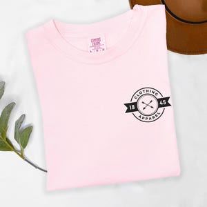 A pink t-shirt with a black logo and text is displayed on a white surface, accompanied by a brown hat and a green plant.