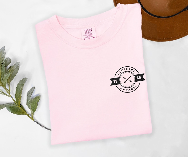 A pink t-shirt with a black logo and text is displayed on a white surface, accompanied by a brown hat and a green plant.