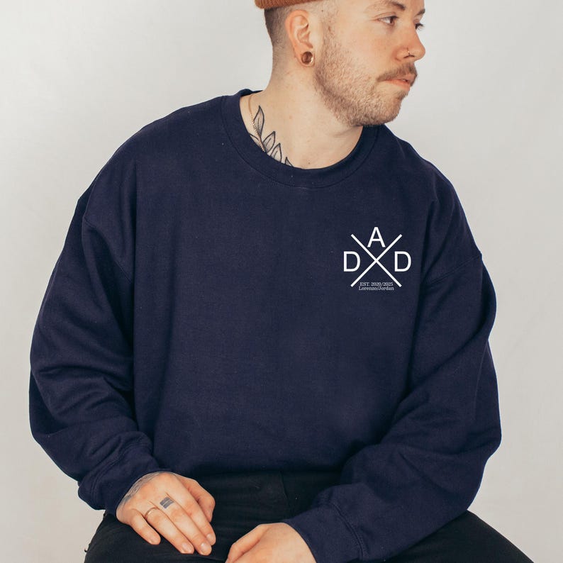 May include: Navy blue sweatshirt with a white graphic on the chest that reads "DAD" in a crossed design. The sweatshirt has a crew neck and long sleeves. The text "EST. 2020" is below the "DAD" graphic.