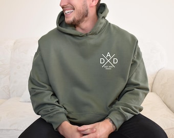 May include: Olive green hoodie with a white graphic on the chest that reads "DAD" in a crossed design, with "EST. 2023 Kansas" below. The hoodie has a hood and is made of a soft material.