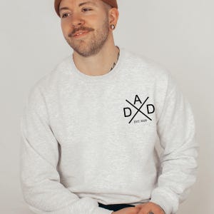 May include: A light gray sweatshirt with the word "DAD" in a crossed design, with the text "EST. 2026" below. The sweatshirt is a crew neck style.
