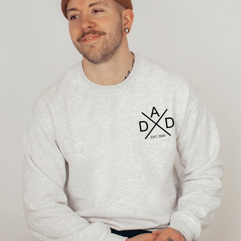 May include: A light gray sweatshirt with the word "DAD" in a crossed design, with the text "EST. 2026" below. The sweatshirt is a crew neck style.