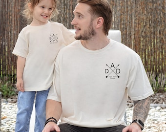 May include: Two cream-colored t-shirts, one worn by a child and the other by an adult. Both shirts feature a black graphic design. The child's shirt has the text "A B B Y" with arrows. The adult's shirt has the text "DAD" with arrows and "Est. 2023 Emma".