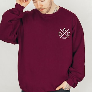 May include: A maroon sweatshirt with the word "DAD" in a white, stylized design on the chest. The design includes the letters "A", "D", and "D" in a cross formation, with "EST. 2026 Carson" below.