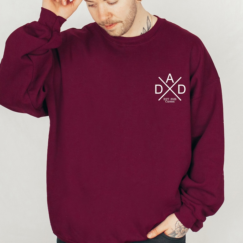 May include: A maroon sweatshirt with the word "DAD" in a white, stylized design on the chest. The design includes the letters "A", "D", and "D" in a cross formation, with "EST. 2026 Carson" below.