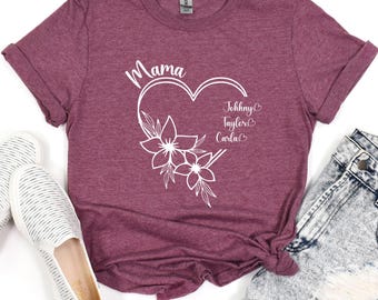May include: Burgundy t-shirt with a white heart design, the word "Mama" above the heart, and the names "Johnny", "Taylor", and "Carla" inside. The heart is decorated with white floral accents. The shirt is paired with denim shorts and white shoes.