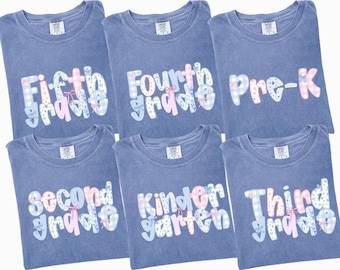 May include: Six blue t-shirts with grade level text in a decorative font. The text includes "Fifth Grade", "Fourth Grade", "Pre-K", "Second Grade", "Kindergarten", and "Third Grade". The text is in white, pink, and blue with floral and gingham patterns.