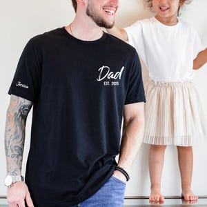 May include: Black t-shirt with the word "Dad" in a script font and the year "2025" printed on the front. The sleeve has the name "Jessica" embroidered on it. A child in a white shirt and beige skirt stands next to the person wearing the shirt.