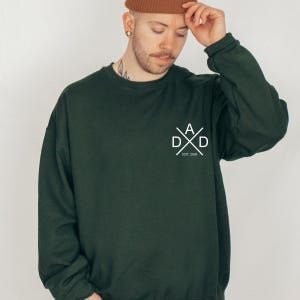 May include: Dark green sweatshirt with a white graphic that reads "DAD" in a stylized design, with "EST. 2026" below. The sweatshirt is a solid color and has a relaxed fit.