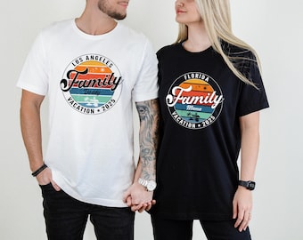 May include: Two short-sleeved t-shirts, one white and one black, featuring a circular design with the words "Family Vacation 2025." The white shirt says "Los Angeles Daddy," and the black shirt says "Florida Mama." The design includes a sunset and palm trees.