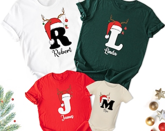 May include: Four Christmas-themed t-shirts and a onesie. The shirts are white, green, and red, and the onesie is beige. Each garment features a letter, name, and a Santa hat design with antlers. The names are Robert, Linda, James, and Mia.
