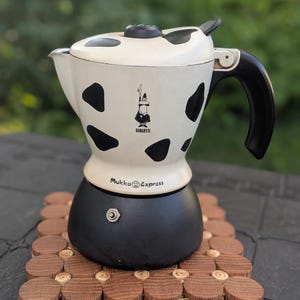 May include: A white and black espresso maker with a cow-like pattern, black handle, and the Bialetti logo. The espresso maker sits on a wooden trivet. The words "Mukka Express" are printed on the front.