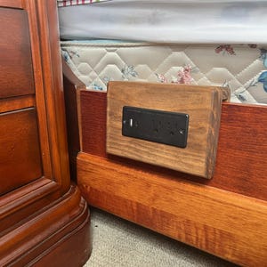 Puede incluir: Una estación de carga de madera para mesita de noche con una toma de corriente negra y un puerto USB. La estación está montada en un marco de cama de madera, junto a una mesita de noche de madera. La cama tiene un colchón con estampado floral.